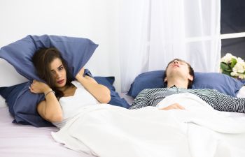 A snoring man sleeping next to an upset woman covering her ears with a pillow.