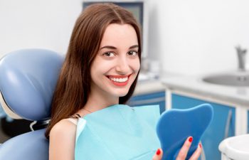 A smiling woman sitting in a dentist's chair holding a mirror.