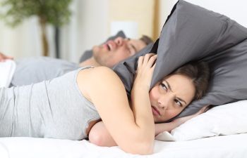 A snoring man sleeping next to an awake woman covering her ears with a pillow.