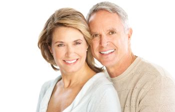 A man and woman smile at the camera in a bright setting. The man has short gray hair and wears a beige sweater, while the woman has shoulder-length light brown hair and wears a white top.