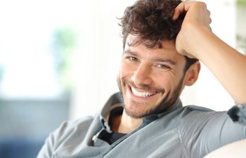 A man with curly hair and a beard sits indoors, smiling at the camera with his hand resting on his head.