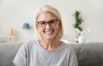 A smiling older woman with short blonde hair and glasses sitting on a couch in a bright living room.