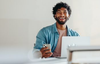 A cheerful man with dark curly hair and a beard, wearing a denim shirt, laughs while looking at the camera, holding a smartphone and sitting at a desk with a laptop.