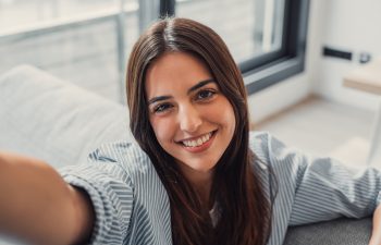 A young woman with long dark hair, wearing a striped shirt, takes a selfie while smiling brightly at the camera. She is indoors on a couch, with a bright window in the background.