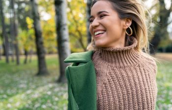 A person wearing a brown knit sweater and green coat stands outdoors, smiling, with blurred trees and grass in the background.