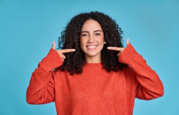 A smiling young woman with curly dark hair, wearing a red knitted sweater, pointing both index fingers at her white teeth against a solid blue background.