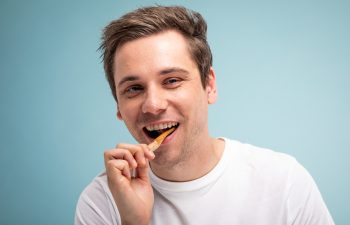 A young man in a white t-shirt looking at the camera and brushing his teeth with a wooden toothbrush against a light blue background.