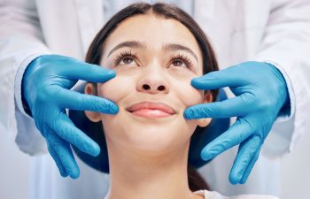 A medical professional in blue gloves gently touching the face of a smiling woman during a skin consultation or aesthetic procedure.